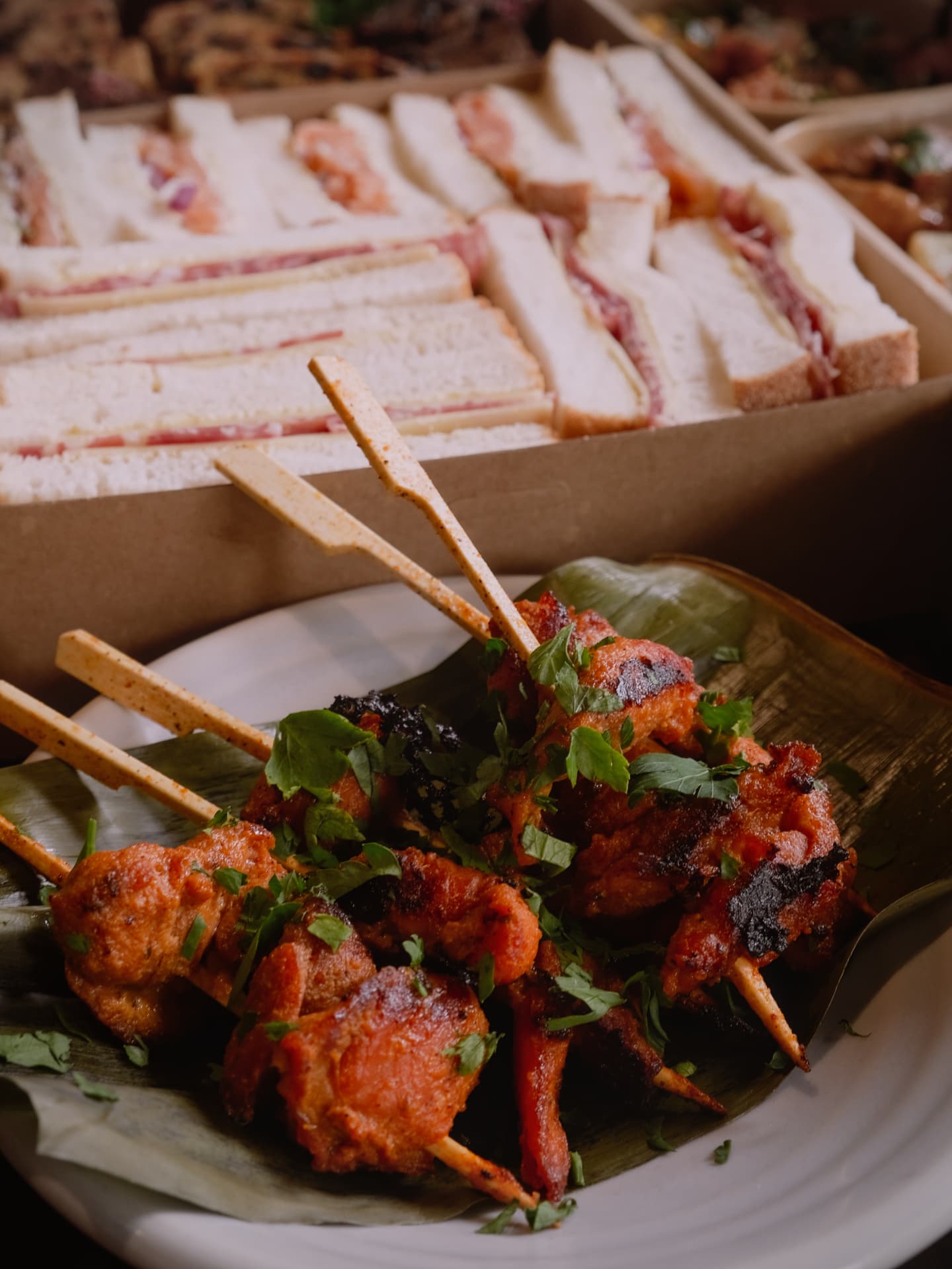 Tandoori chicken skewers served on a banana leaf with fresh coriander, next to a box of sandwiches.