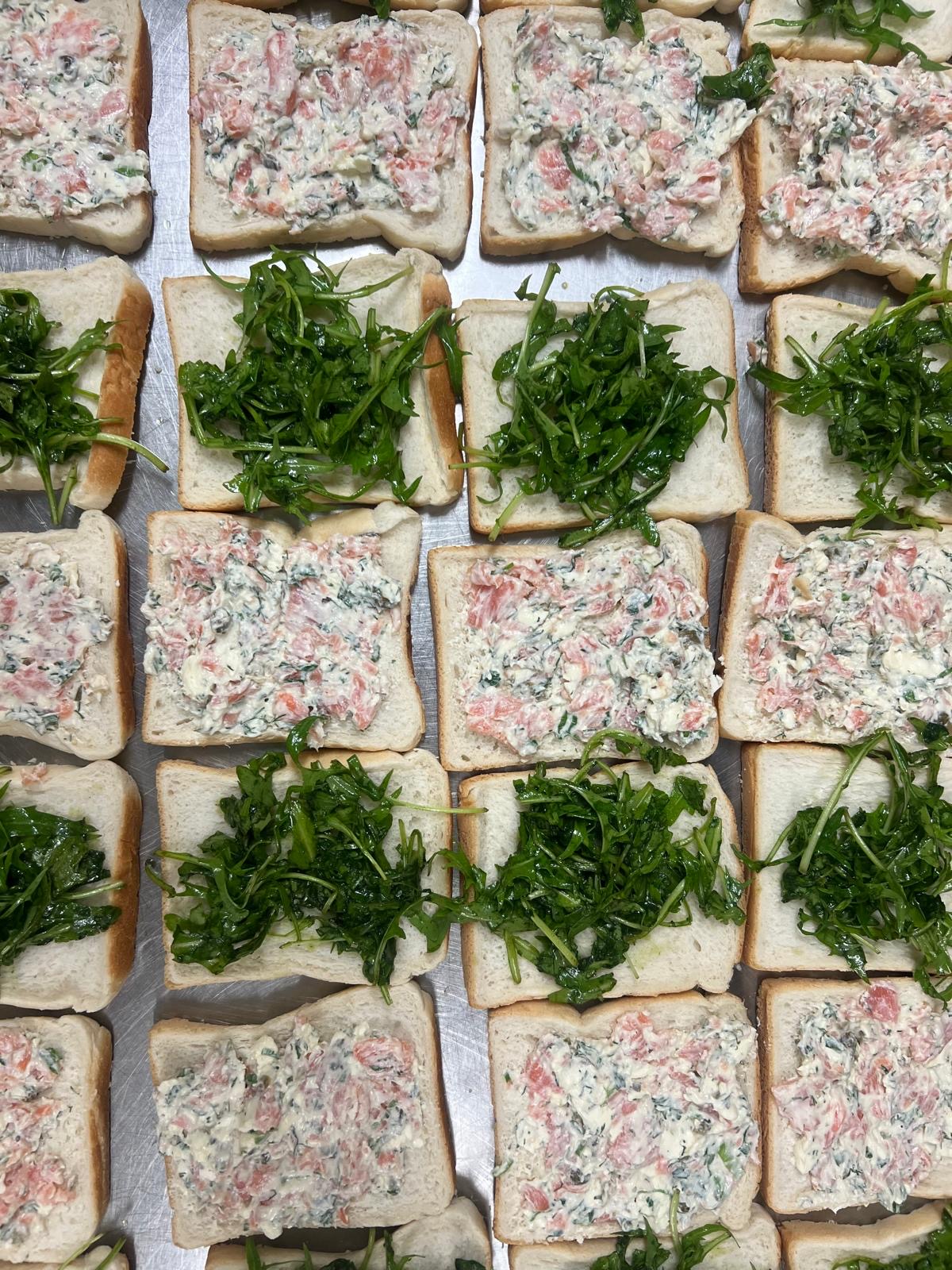 Open faced smoked salmon sandwiches with cream cheese and rocket on a prep tray.