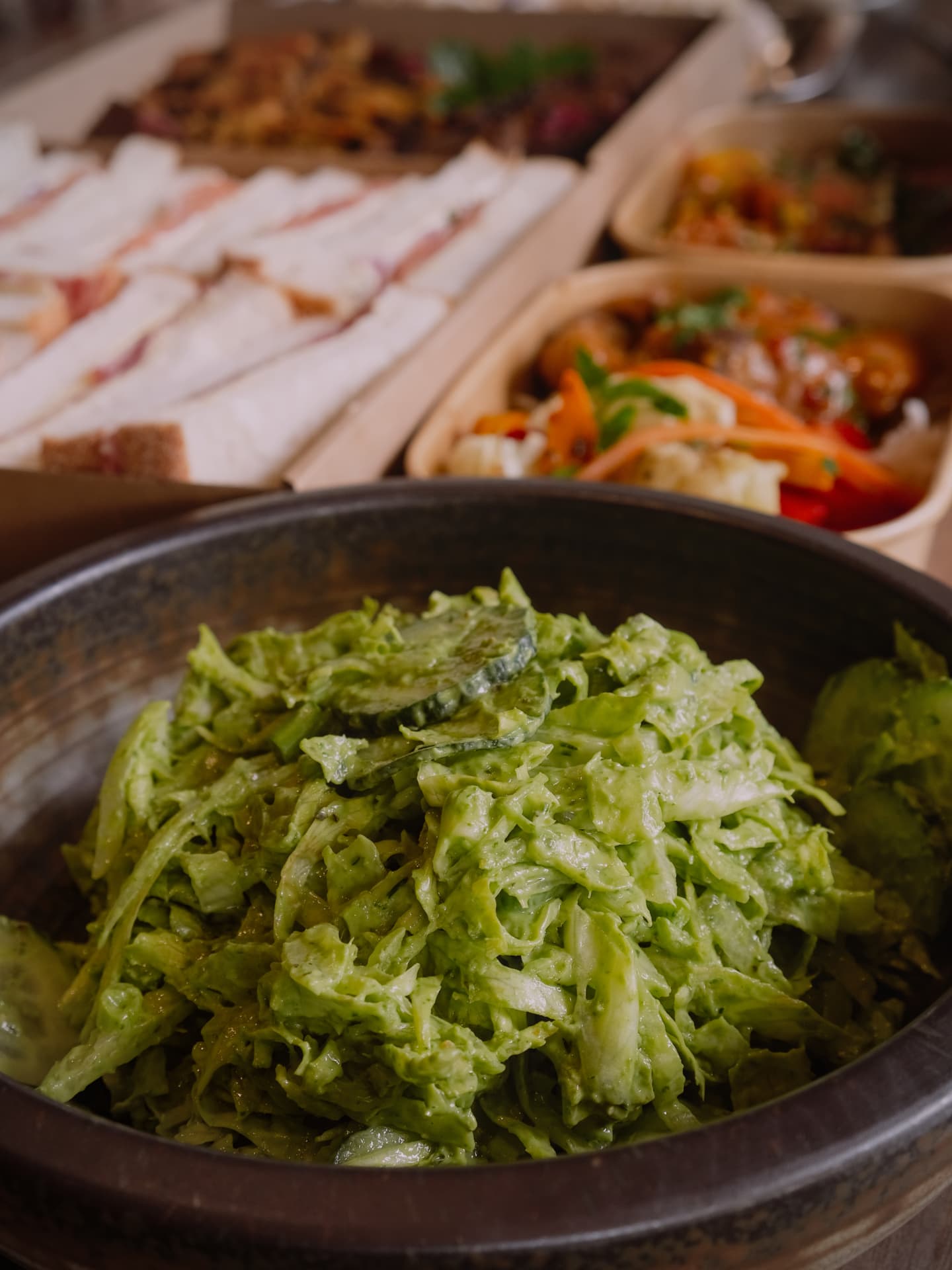 Fresh green slaw in a serving bowl with sandwiches and salads behind it.